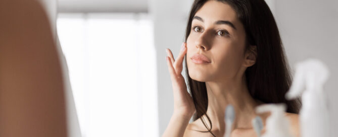 Woman gently touching her cheek while looking in the mirror, representing natural skin appearance and thoughtful Botox decision-making.